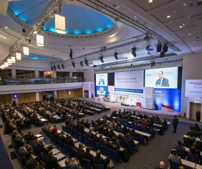 A bustling conference hall with a backdrop of a UK map, attendees in navy suits holding gold‑accented name badges, and a central stage where a speaker presents a property development slide.